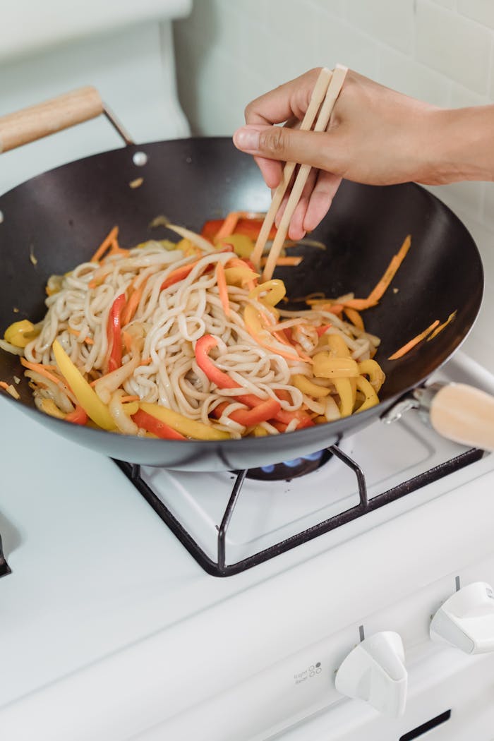 Services Hand using chopsticks to stir fry noodles and bell peppers in a wok on a stove.